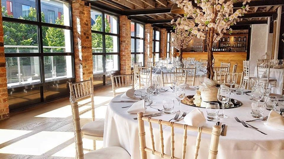 Tables set for a wedding with floor to ceiling windows and a bar in the background